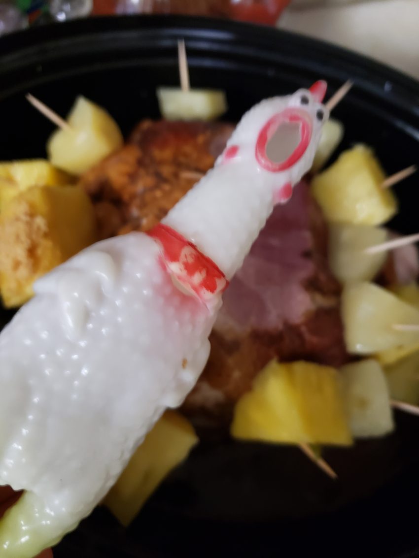 Chick-o, my rubber chicken, is in the foreground with a ham in the background full of toothpicked pineapple chunks in a crockpot. Someone has cut off a piece of the ham